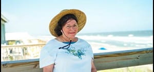 Carol F headshot of her standing on a deck in straw sunhat with beach and ocean in the background
